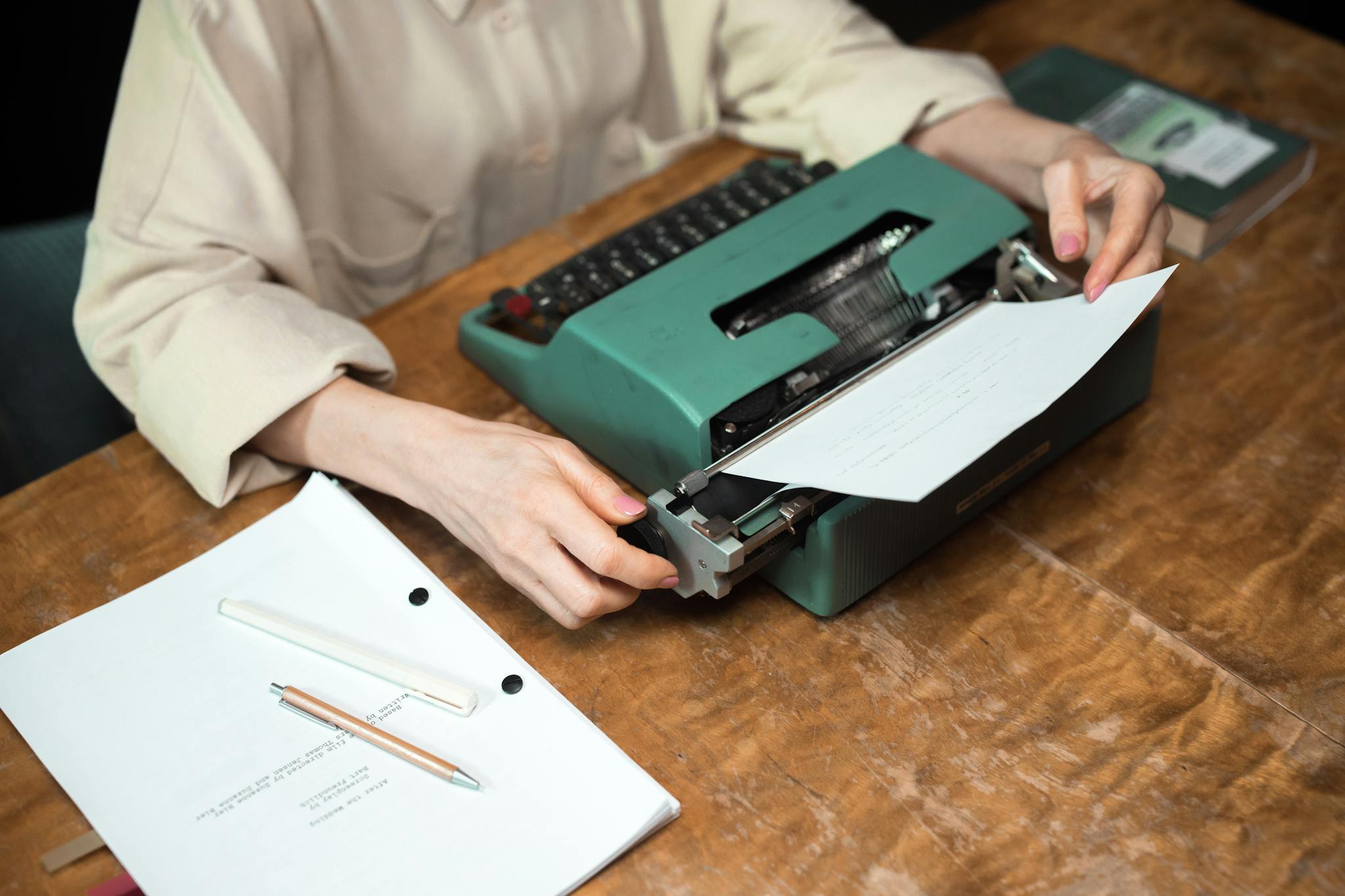 Close-up of hands using a vintage typewriter, with paper and script on a wooden desk.