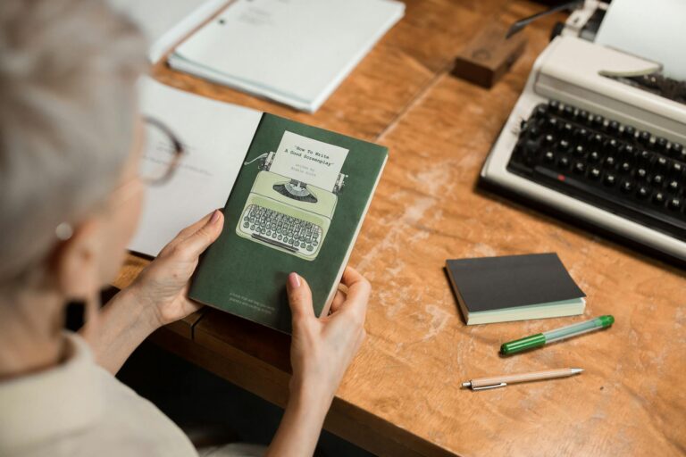 Elderly woman holding a book at a wooden desk with a classic typewriter, creating a retro office vibe.
