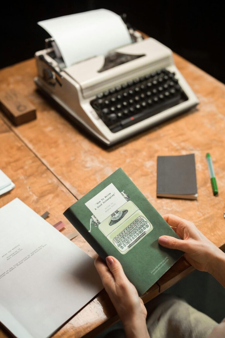 Hands hold a vintage book in front of an old typewriter on a wooden desk.